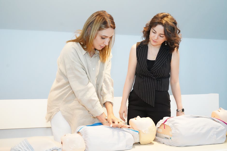 Two women practice CPR on adult manikins during a first aid training session in a bright room.