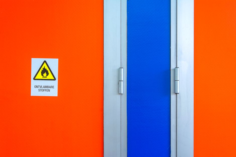 Orange wall with a blue vertical panel/door and a safety label showing a flame icon and the text 'Ontvlambare stoffen' (Flammable materials).