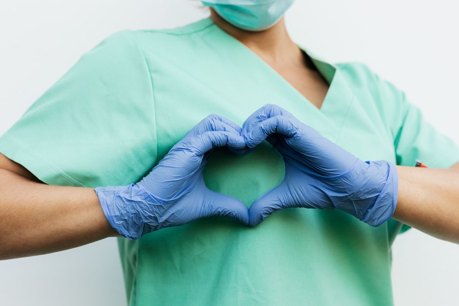 Healthcare worker in green scrubs wearing a mask forms a heart shape with blue gloves to symbolize care.