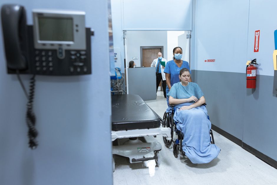 Hospital hallway: a patient in a wheelchair covered with a blue blanket is being pushed by a nurse in blue scrubs, with another staff member in the background.