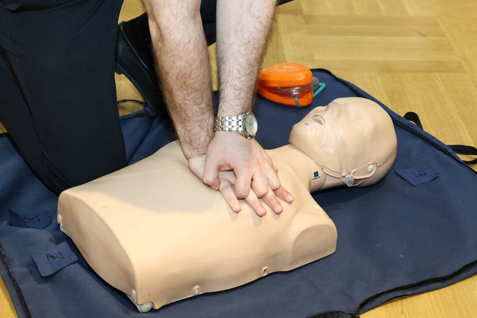 Person performing chest compressions on a CPR manikin during training on a blue mat with an orange mask nearby.