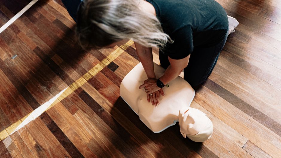 Person performing chest compressions on a CPR manikin during a training session on a wooden gym floor, hands interlocked and shoulders above the chest.