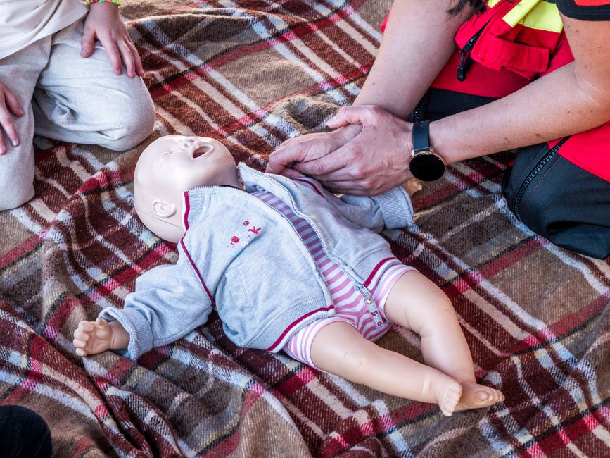 Infant CPR training doll lying on a plaid blanket as an adult demonstrates chest compressions during a first-aid class.
