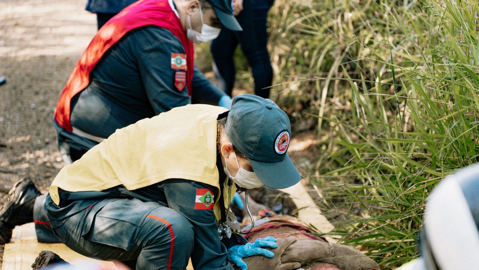 Rescue workers in uniforms and masks kneel by an injured person lying on the ground outdoors near tall grasses, providing aid.