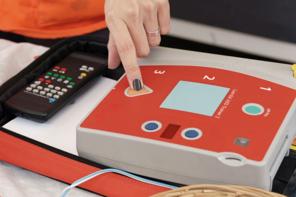 - Aspire First Aid Guide Training Corp Person pressing a button on a red AED trainer device, with a remote control nearby.