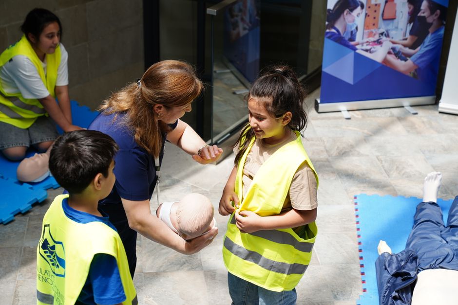 Instructor demonstrates infant bandaging to children in high-visibility vests during a first aid training session.